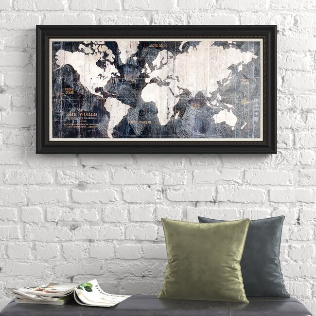 A 24\“ x 36\“ framed old-world style map features a vintage design with a black frame, displayed on a textured white brick wall. Two pillows and magazines are below the map for added decor.