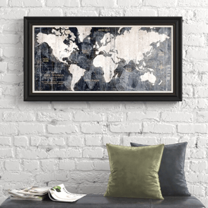 A 24\“ x 36\“ framed old-world style map features a vintage design with a black frame, displayed on a textured white brick wall. Two pillows and magazines are below the map for added decor.