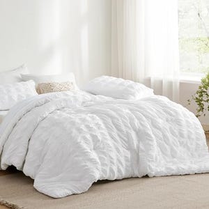 A white seersucker comforter set is displayed on a bed, with textured stripes providing a ruffled look. The set includes matching pillow shams behind a decorative pillow.