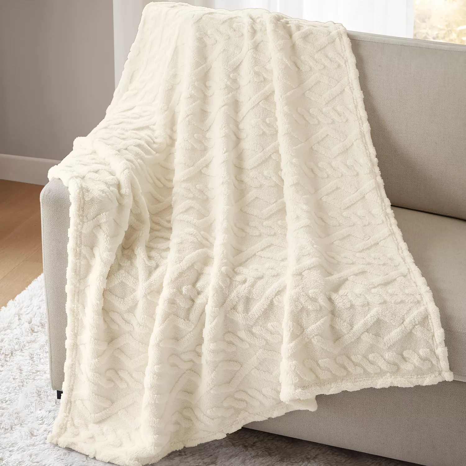A plush, ivory throw blanket with a textured geometric pattern is draped over a light gray sofa.