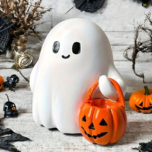 A small, cute ghost figurine holding an orange pumpkin-style bucket, surrounded by Halloween decorations like black spiders, bats, and mini pumpkins on a wooden surface.