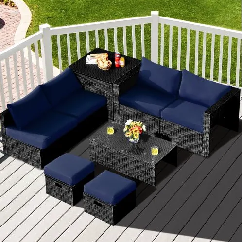 Outdoor patio set with dark wicker furniture, blue cushions, and a glass-top coffee table. Includes an L-shaped sofa, two stools, and a multi-level corner piece with snacks and drinks displayed.