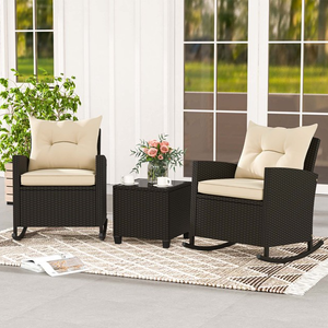 A patio set with two wicker rocking chairs featuring beige cushions and a matching side table. A decorative rug, potted plant, and a vase with flowers complement the setup.