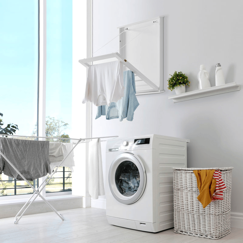 Laundry room with a front-loading washing machine, wall-mounted drying rack with clothes, foldable drying rack with more clothes, a wicker laundry basket, and a shelf with laundry supplies and a plant.