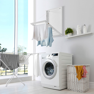 Laundry room with a front-loading washing machine, wall-mounted drying rack with clothes, foldable drying rack with more clothes, a wicker laundry basket, and a shelf with laundry supplies and a plant.