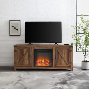 A wooden TV stand with a barn door design features a central electric fireplace insert, flanked by two cabinets with diagonal crossbar accents. It stands against a white brick wall and supports a flat-screen TV on top.