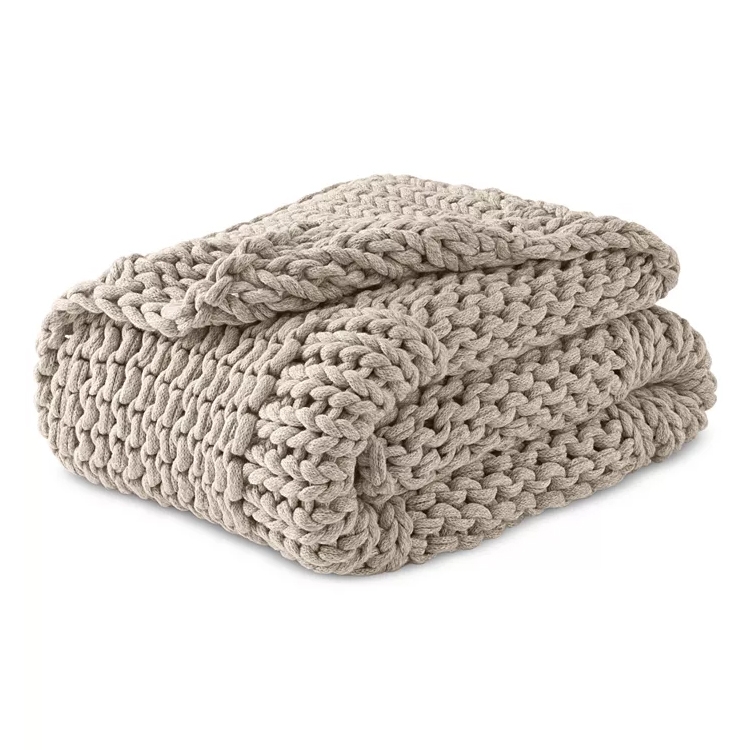 A beige chunky knit throw blanket with large, woven stitches, folded neatly. The thick texture suggests a cozy, warm feel.