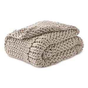 A beige chunky knit throw blanket with large, woven stitches, folded neatly. The thick texture suggests a cozy, warm feel.