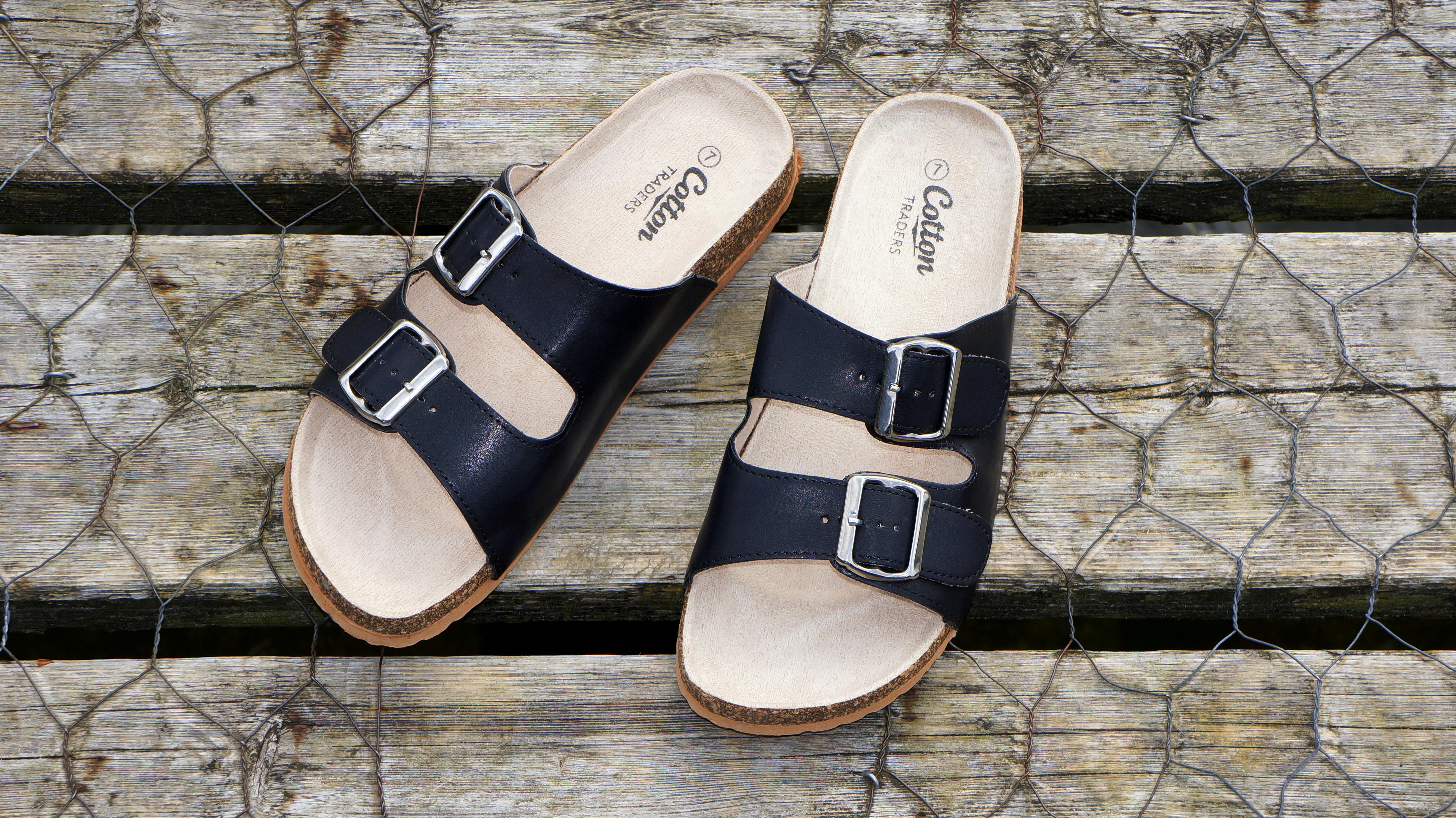 A pair of black two-strap sandals with buckle closures and beige footbeds, displayed on a wooden surface.