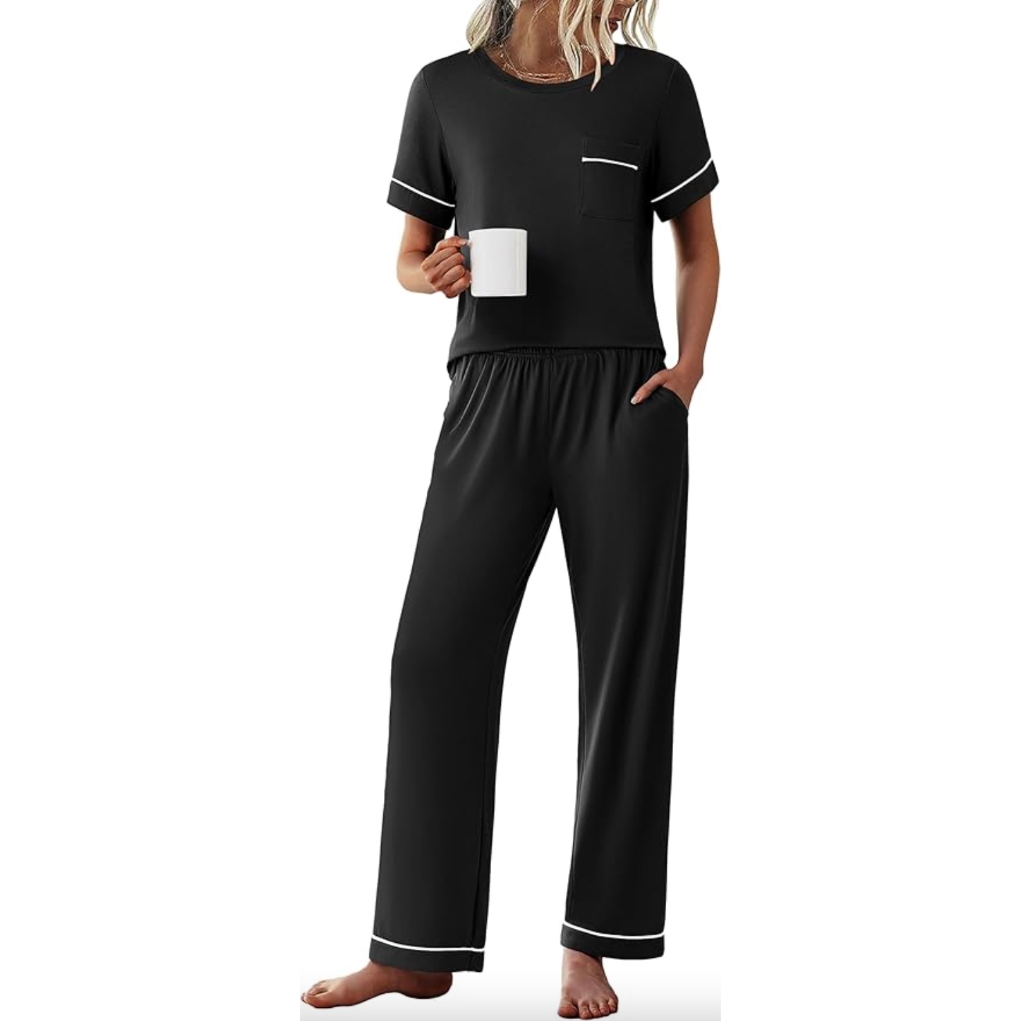 Black two-piece loungewear set with a short-sleeve top featuring a chest pocket and white trim on the sleeves, paired with loose-fitting pants that have an elastic waistband and white trim at the hem. The person is holding a white mug and standing barefoot.