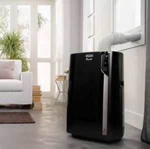 A portable air conditioner with a flexible exhaust hose attached to a window is placed in a modern living room near a couch and a stack of books.
