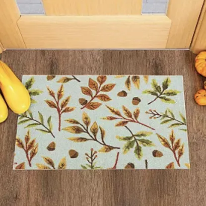 Autumn-themed doormat with a leaf and acorn pattern, placed in front of a wooden door. Yellow rain boots are on the left, and small pumpkins are on the right.