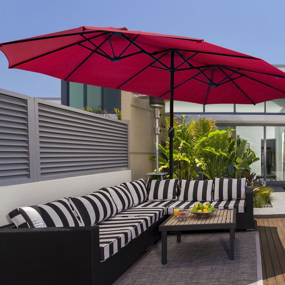 A patio setup with a black and white striped outdoor sectional sofa, a wooden coffee table with fruit and drinks, and a large red patio umbrella providing shade. The setting includes greenery and modern architectural elements in the background.