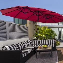 A patio setup with a black and white striped outdoor sectional sofa, a wooden coffee table with fruit and drinks, and a large red patio umbrella providing shade. The setting includes greenery and modern architectural elements in the background.