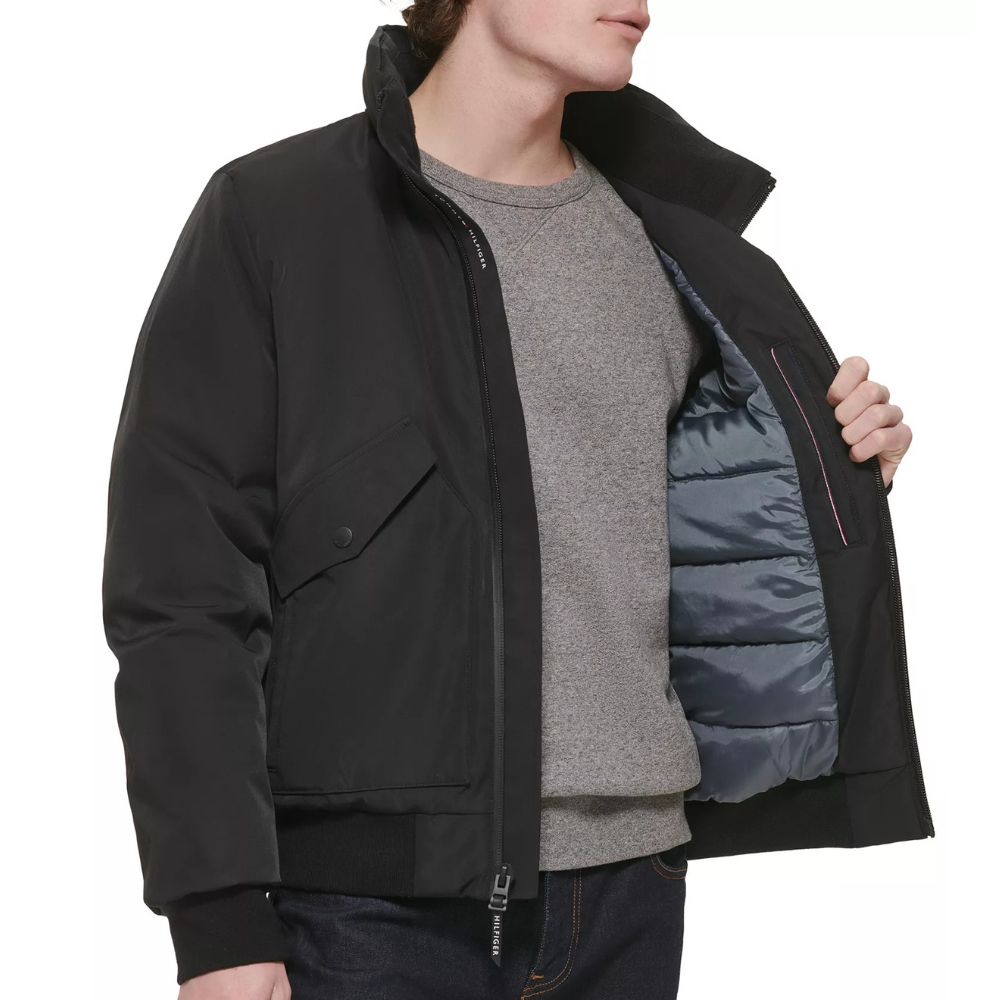A man models a black jacket with a quilted inner lining, showcasing its insulation. He wears a gray sweater underneath, and the jacket features a flap pocket and zipper closure.