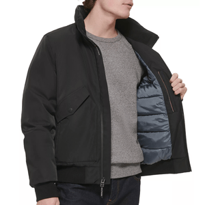 A man models a black jacket with a quilted inner lining, showcasing its insulation. He wears a gray sweater underneath, and the jacket features a flap pocket and zipper closure.