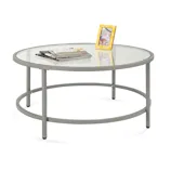 Round coffee table with a glass top and metal frame, featuring a photo frame and magazines on the surface.