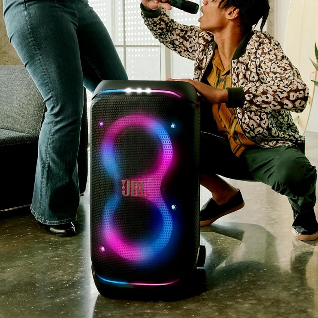 A person is singing into a microphone next to a large JBL portable speaker with colorful LED lights.