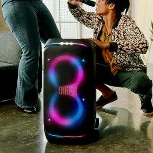 A person is singing into a microphone next to a large JBL portable speaker with colorful LED lights.