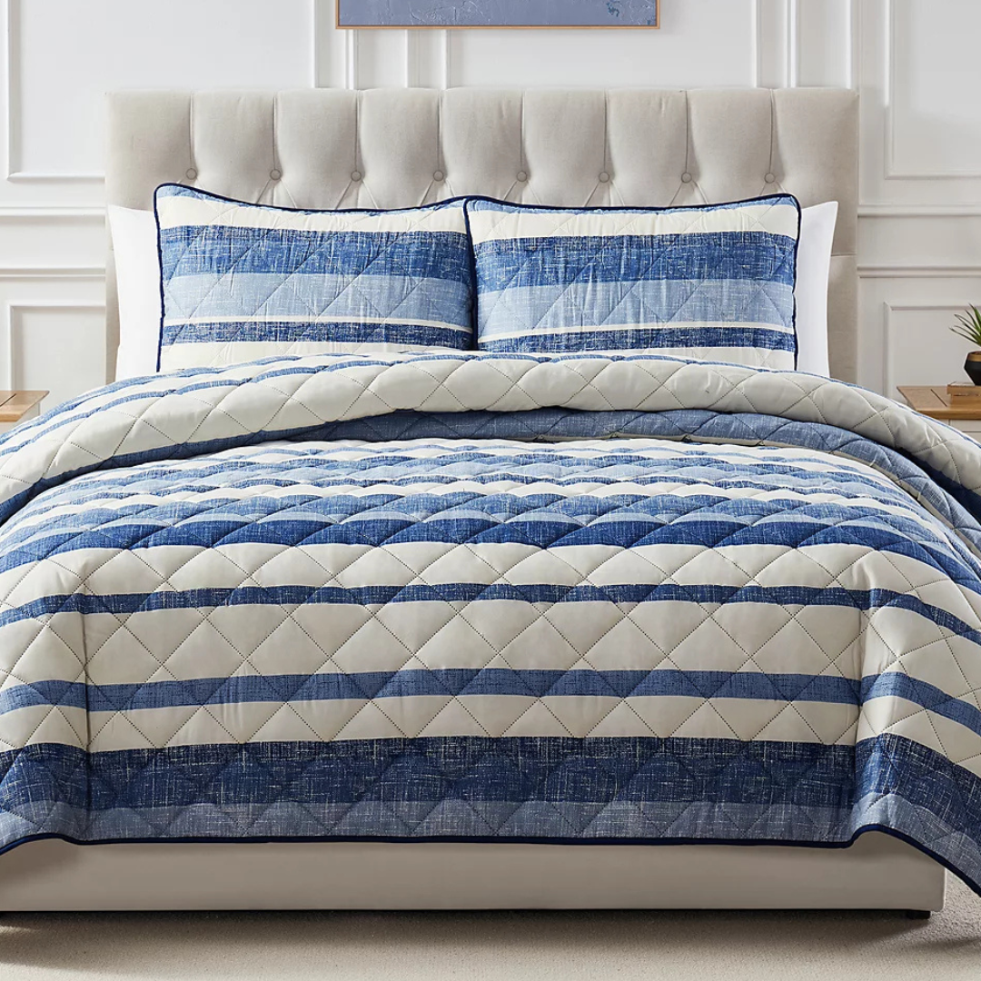 A blue and white striped quilt with matching pillow shams is displayed on a beige tufted headboard bed. The quilt features a diamond stitch pattern.