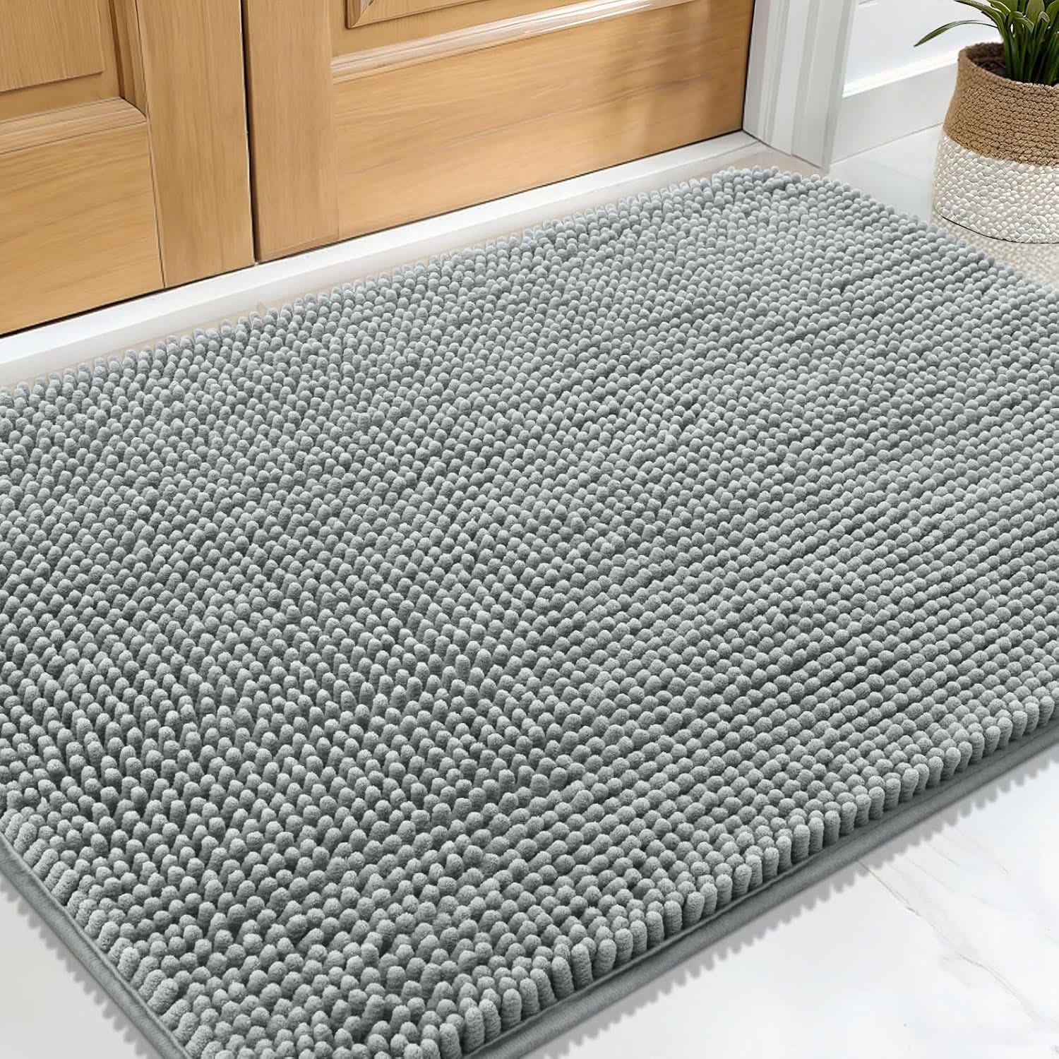 A gray chenille rug with a textured, looped surface is placed in front of a wooden door, accompanied by a potted plant on the side.
