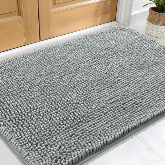 A gray chenille rug with a textured, looped surface is placed in front of a wooden door, accompanied by a potted plant on the side.