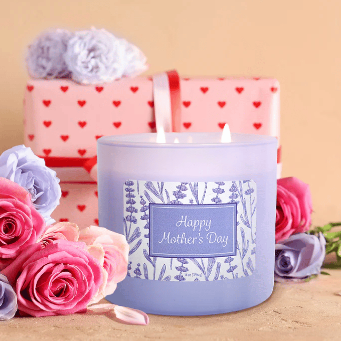 A lavender-scented candle labeled \“Happy Mother's Day\“ is surrounded by pink and purple roses. Behind it, there's a gift wrapped in heart-patterned paper with a red ribbon.