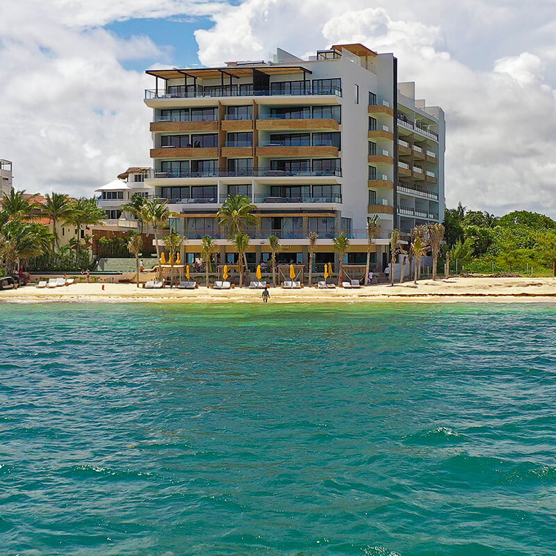 A beachfront hotel with multiple stories features balconies, surrounded by palm trees and sun loungers on the sandy beach, adjacent to clear turquoise waters.