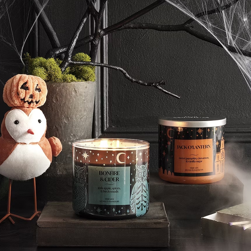 Two scented candles, \“Bonfire & Cider\“ and \“Jack-O-Lantern,\“ are displayed beside a plush penguin with a pumpkin hat. Halloween-inspired decorations complement the scene.