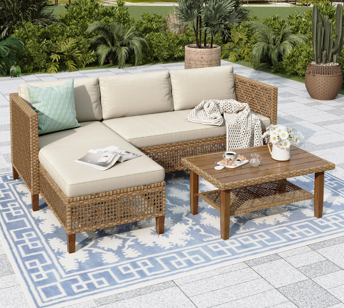 A wicker patio sofa set featuring beige cushions with one teal pillow and a knitted throw, paired with a matching wooden coffee table. The setup includes a blue and white patterned outdoor rug and a white flower vase on the table.