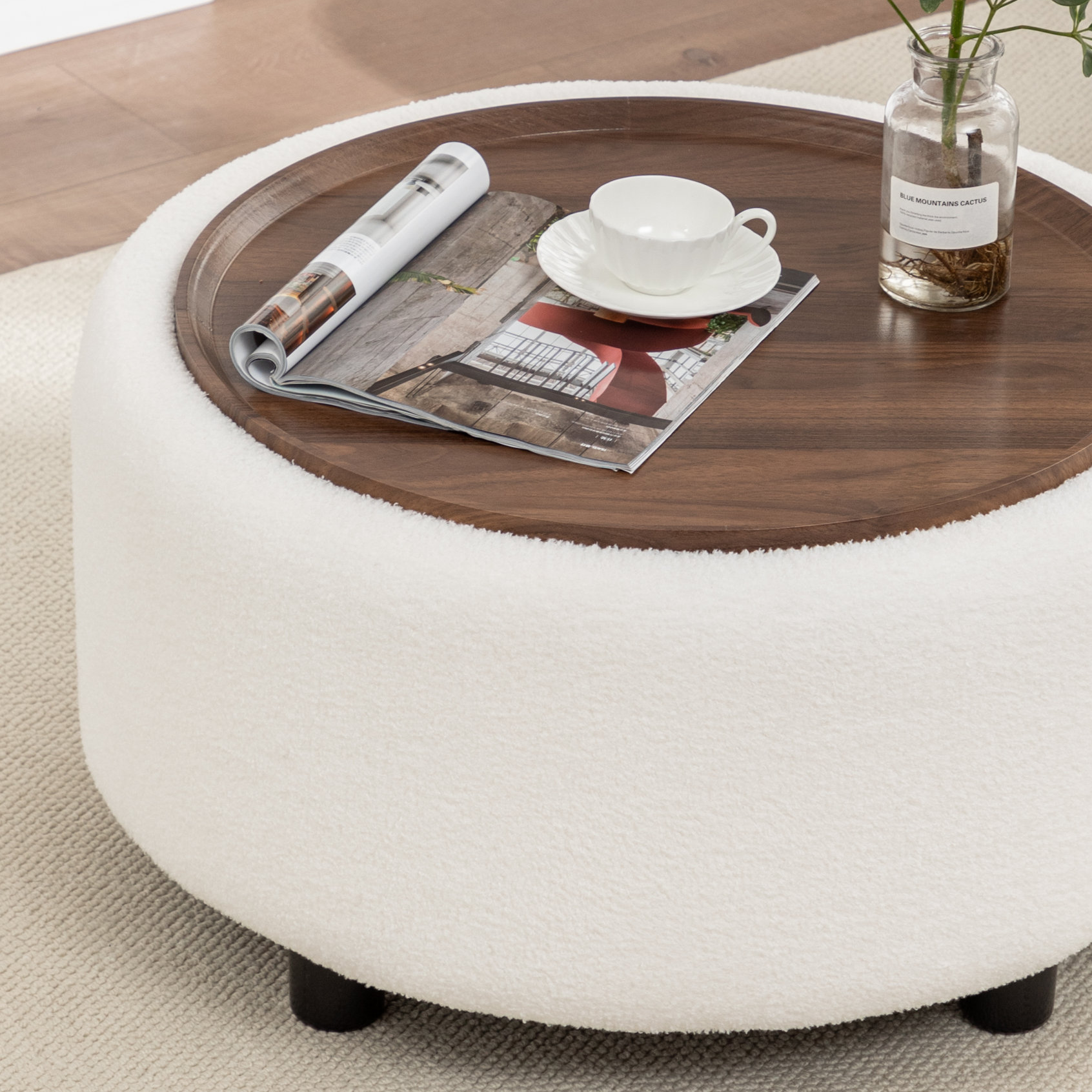 A round, white boucle storage coffee table with a diameter of 28 inches, featuring a wooden top. The table holds a magazine, a white teacup, and a glass vase with a label.