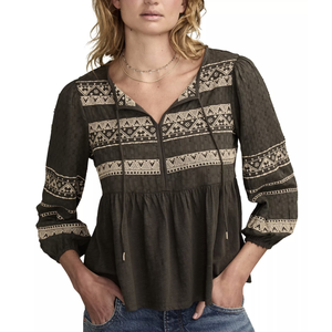 A woman is wearing a dark blouse with beige geometric embroidery patterns and drawstrings on the neckline, paired with blue jeans.