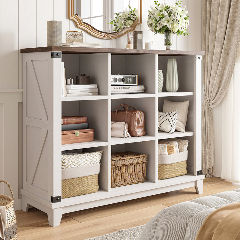 A white storage cabinet with a dark wood top features nine cubbies, housing decorative items such as books, vases, a radio, baskets, and a handbag. A mirror and vase with flowers are on top.