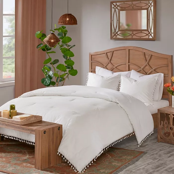 A bedroom setup with a wooden bed frame, white bedding with tassel edges, pendant lights, and a bench at the foot of the bed.