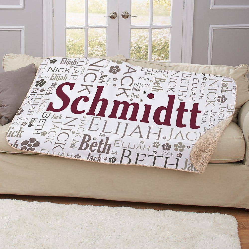 A personalized blanket featuring the name \“Schmidtt\“ prominently, surrounded by additional names like Elijah, Beth, Jack, and Nick, along with paw prints, displayed on a sofa.