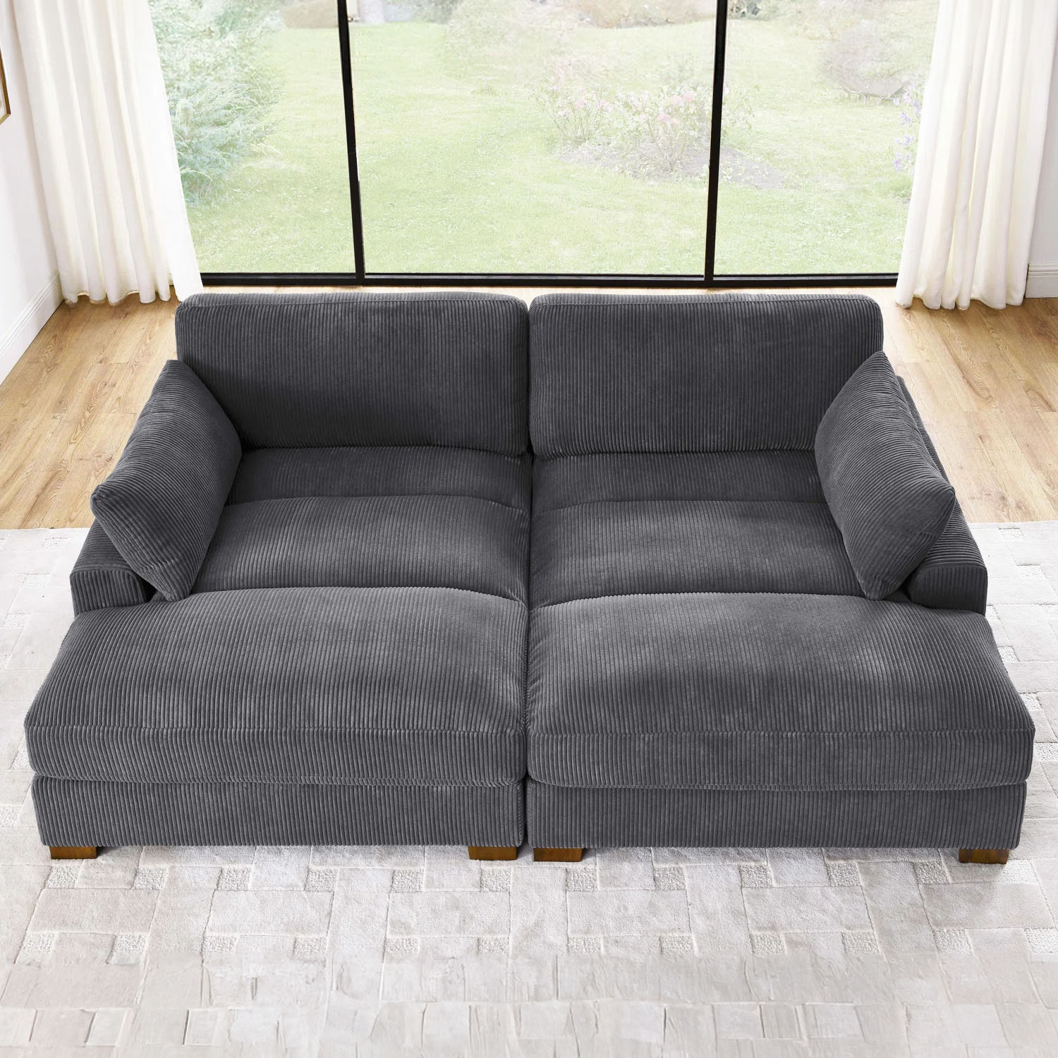 Gray corduroy modular sofa with deep cushions and a contemporary design, placed on a textured white rug in front of large windows with a view of a green yard.