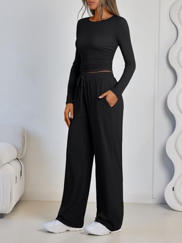 A woman wearing a black long-sleeve crop top paired with black wide-leg pants, standing beside a white sofa and holding her hands in the pants' pockets.