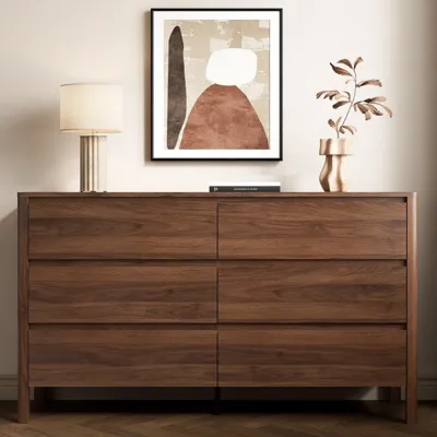 Walnut dresser with two rows of three drawers, topped with a beige lamp and a decorative vase with leaves. Abstract framed art hangs above.