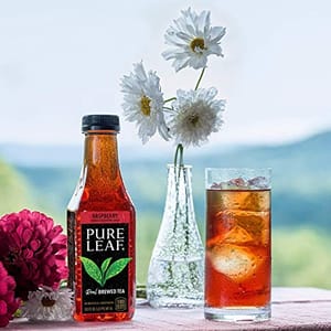 A bottle of Raspberry flavor Pure Leaf Iced Tea is displayed beside a glass filled with the beverage and ice, set against a scenic outdoor backdrop with flowers in a vase.