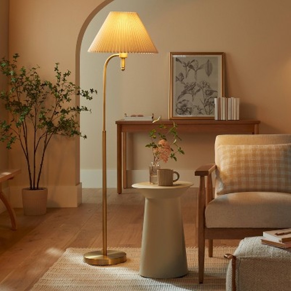 A pleated metal floor lamp with a slender gold stand features a classic pleated beige shade, providing soft ambient lighting. It complements the warm, neutral-toned room decor.