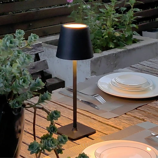 A patio scene with a slim black table lamp, a wooden table set with white dinnerware and silverware, and potted plants in the background.