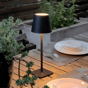 A patio scene with a slim black table lamp, a wooden table set with white dinnerware and silverware, and potted plants in the background.