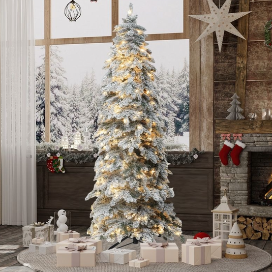 A snowy, pre-lit Christmas tree stands in a cozy, decorated living room with a fireplace, surrounded by wrapped gifts.