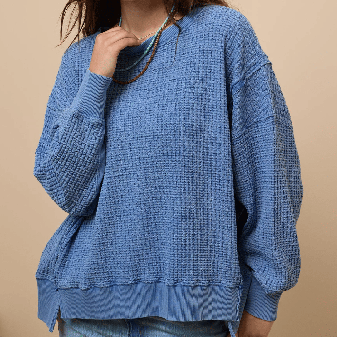 A blue, waffle-knit sweatshirt featuring a loose, oversized fit with drop shoulders and ribbed cuffs and hem. It's worn with a multicolored beaded necklace.
