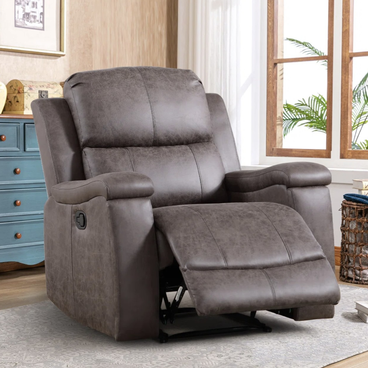 A gray reclining armchair with plush cushions is set in a cozy room with wooden flooring, a patterned rug, a blue cabinet, and a window with green plants in view.
