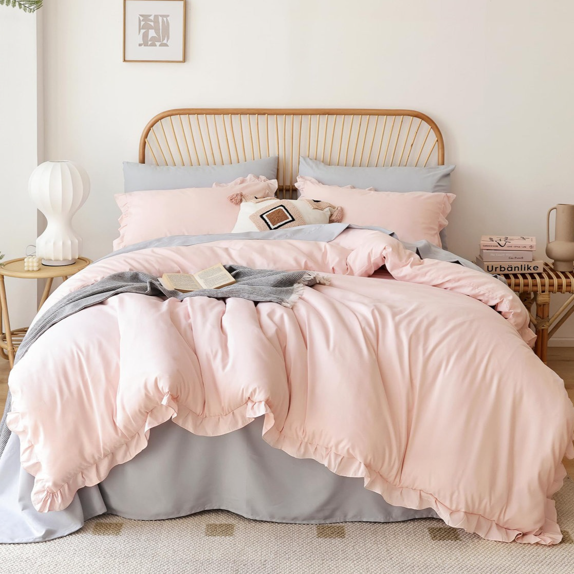 A cozy bedroom features a rattan headboard, soft pink and gray bedding with ruffled edges, a decorative pillow, a book, and a gray throw blanket. A modern white lamp and books are on the bedside table.