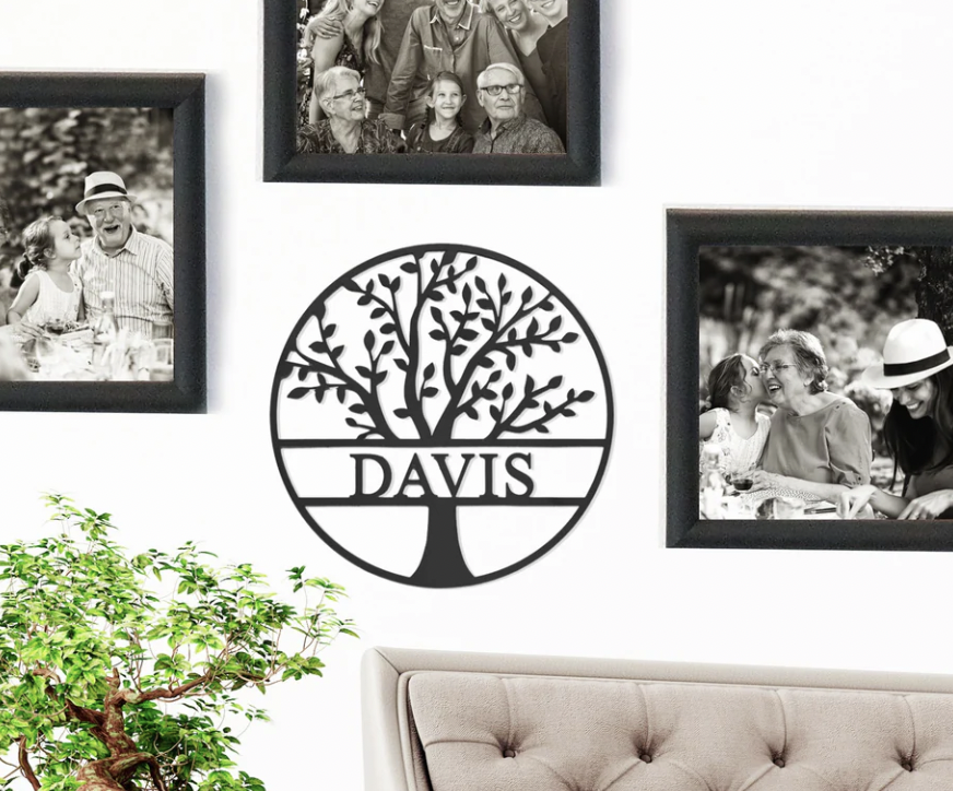A black, circular metal sign featuring a tree design with branches and leaves, and the word \“DAVIS\“ across the center. Framed family photos surround the sign, with a small plant and part of a sofa visible below.