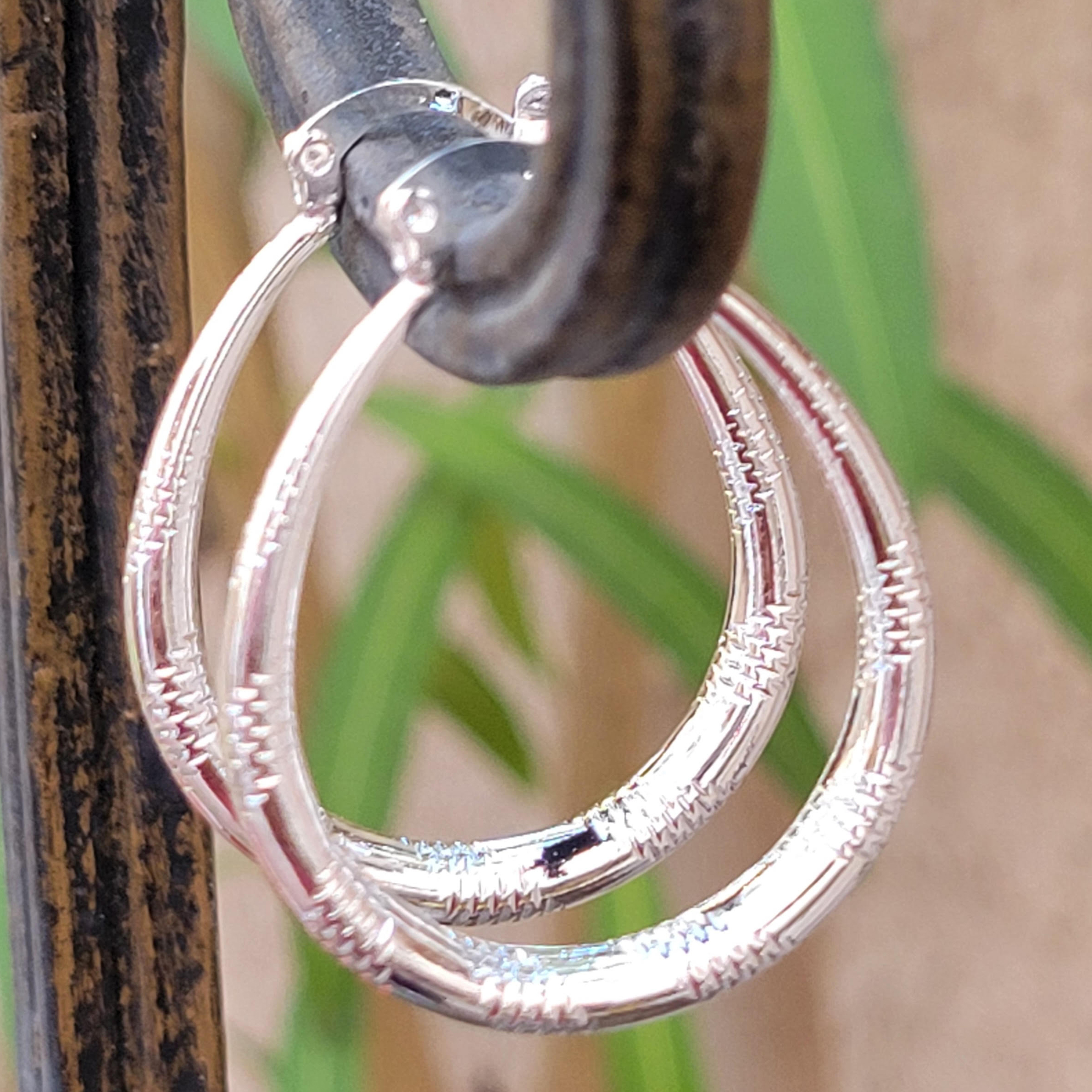 Silver hoop earrings with a textured design are hung on a metal hook. A blurred green plant is visible in the background.