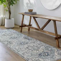 A patterned runner rug is laid on a wooden floor next to a rustic wooden bench with books on top. A potted plant is positioned nearby, close to a window with sheer curtains.