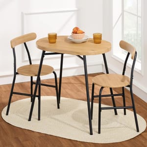 A small round wooden dining set with two chairs on a light oval rug, featuring black metal frames. The table holds two amber glasses and a bowl of peaches.